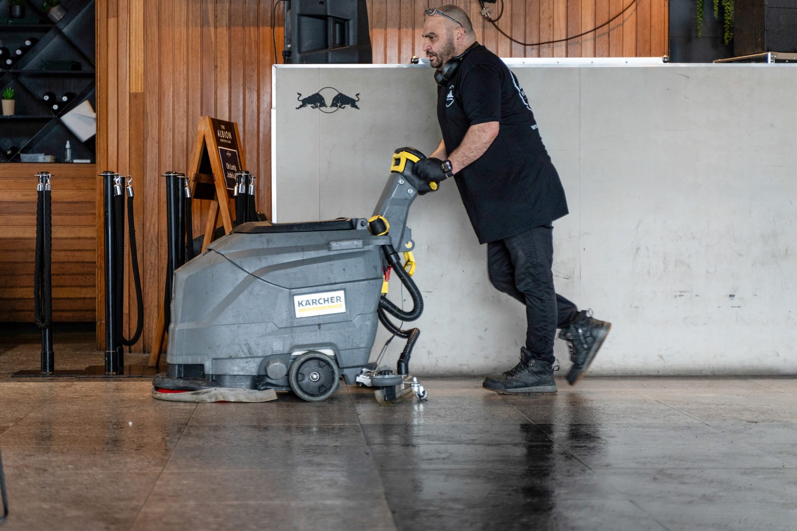A HOZ CLEANING cleaner guiding a ride-behind floor scrubber across a venue floor.